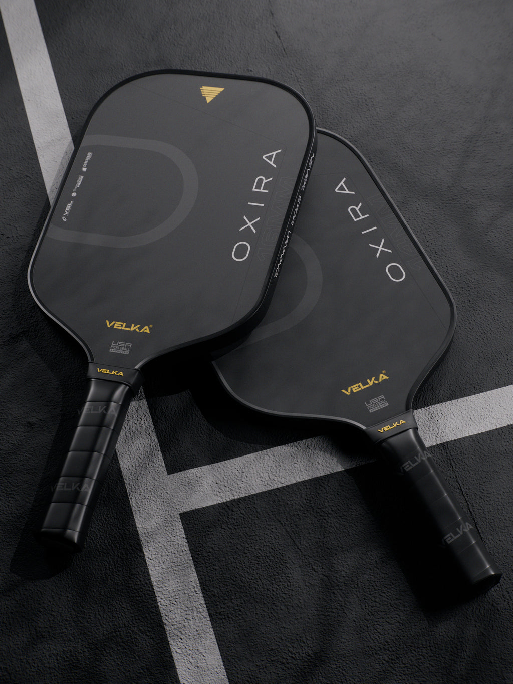 Two black pickleball paddles with 'OXIRA' and 'Velka' branding on a dark pickleball court.