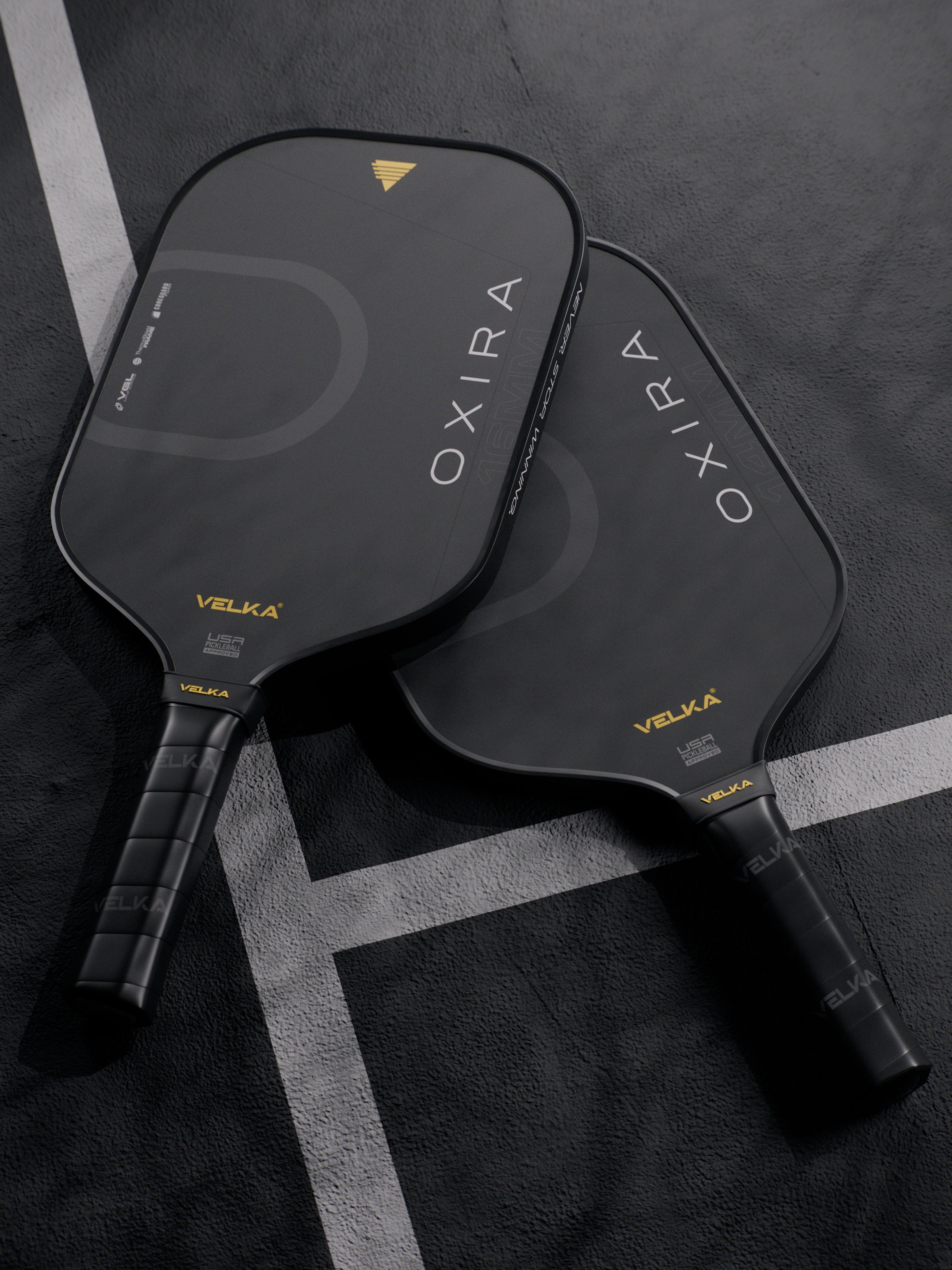 Two black pickleball paddles with 'OXIRA' and 'Velka' branding on a dark pickleball court.