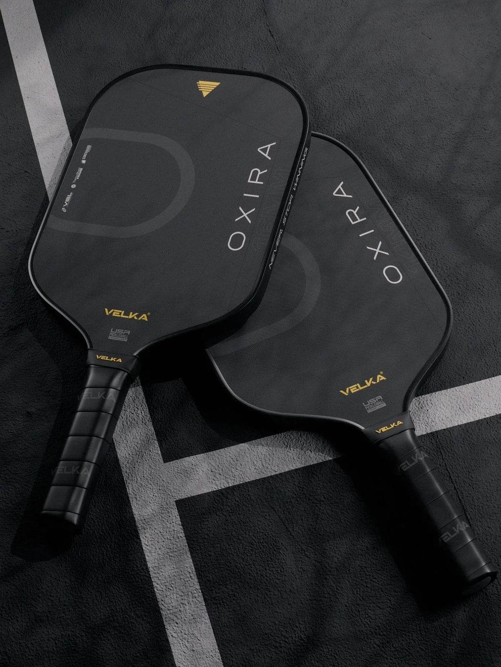 Two black pickleball paddles with 'OXIRA' and 'Velka' branding on a dark pickleball court.