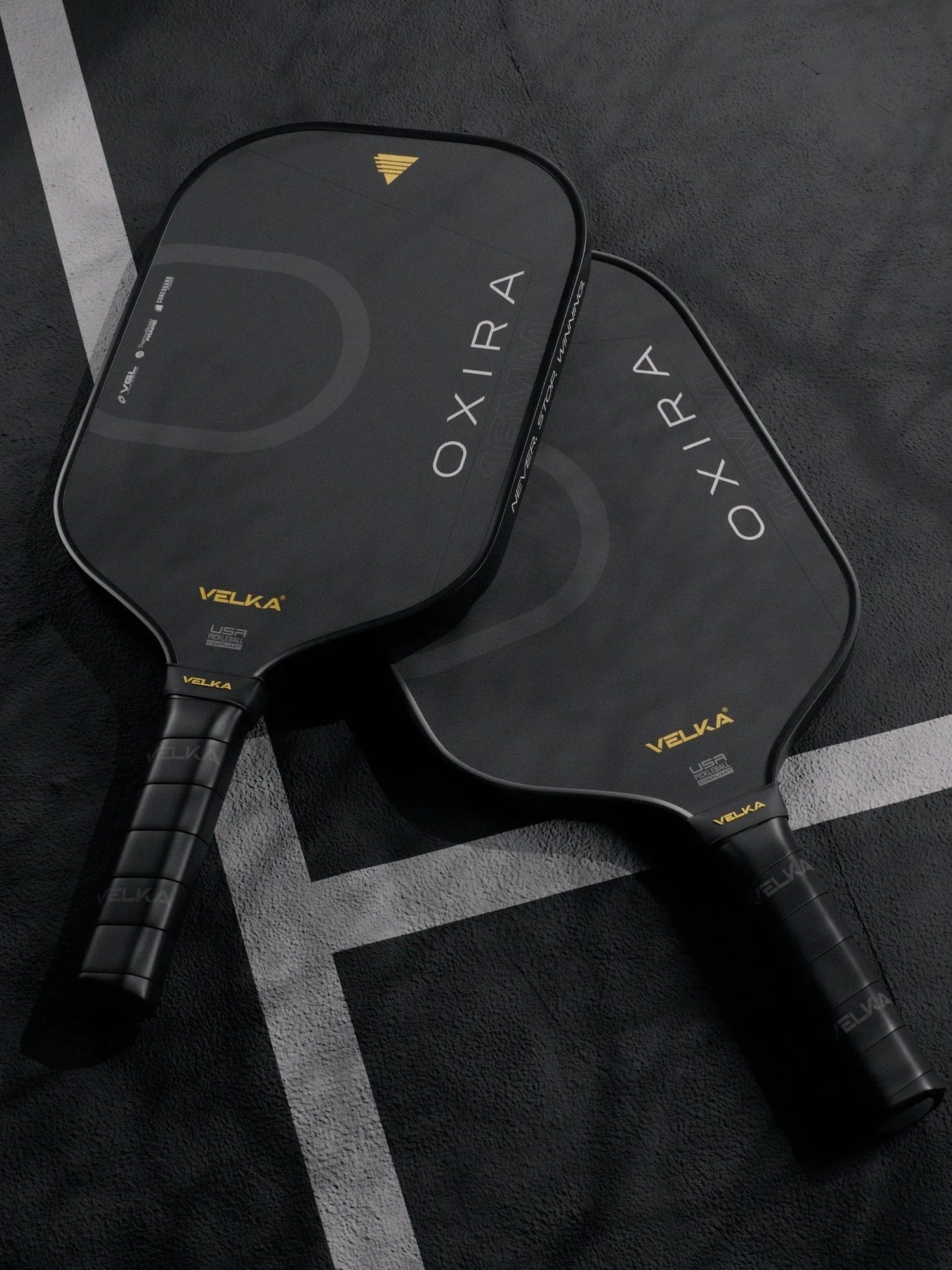 Two black pickleball paddles with 'OXIRA' and 'Velka' branding on a dark pickleball court.